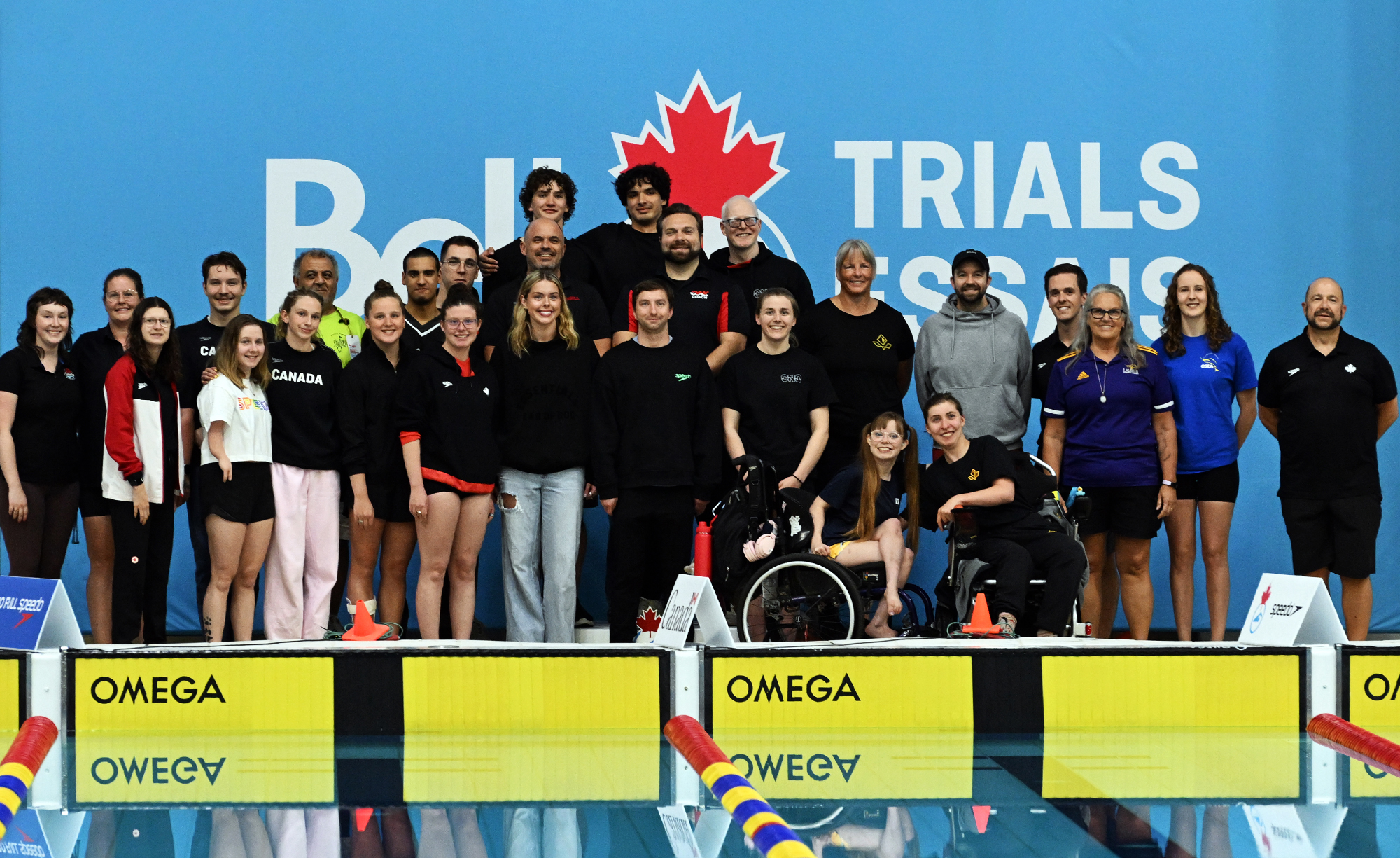 Group photo of the Para swimming squad and staff for Swimming Canada standing on the pool deck in front of a blue “Bell Trials” backdrop, with athletes and coaches smiling together.