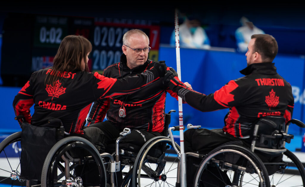 Canadian wheelchair curlers Ina Forrest, Mark Ideson, and Jon Thurston fist bump during a game at the Beijing 2022 Paralympic Winter Games. All three athletes wear black and red Team Canada jackets with their names on the back and are seated in wheelchairs on the curling rink.