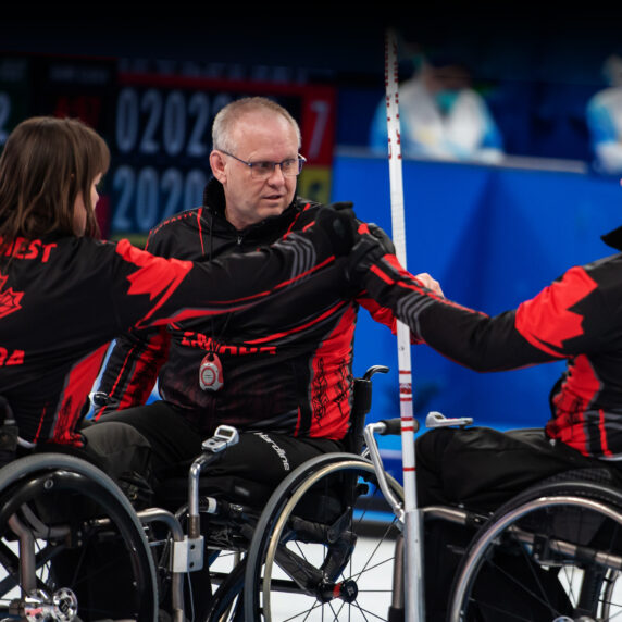 Canadian wheelchair curlers Ina Forrest, Mark Ideson, and Jon Thurston fist bump during a game at the Beijing 2022 Paralympic Winter Games. All three athletes wear black and red Team Canada jackets with their names on the back and are seated in wheelchairs on the curling rink.