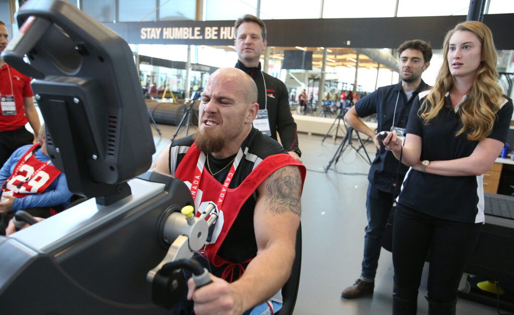 A determined Para athlete pushes hard on an arm ergometer during a fitness test, wearing a red Canada bib numbered 30. Three evaluators, two men and one woman with a stopwatch, closely observe his performance in a gym with the words "STAY HUMBLE. BE HUMBLE." displayed in the background.