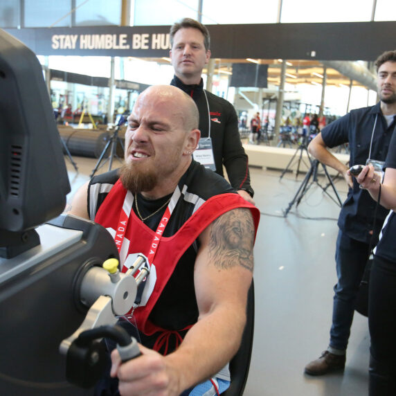 A determined Para athlete pushes hard on an arm ergometer during a fitness test, wearing a red Canada bib numbered 30. Three evaluators, two men and one woman with a stopwatch, closely observe his performance in a gym with the words "STAY HUMBLE. BE HUMBLE." displayed in the background.