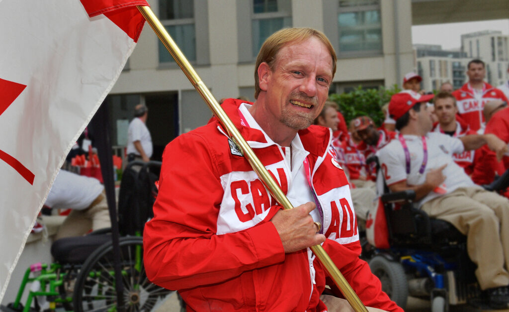 Garett Hickling smiles while proudly holding the Canadian flag, wearing a red and white Team Canada jacket, surrounded by teammates at a Paralympic event.