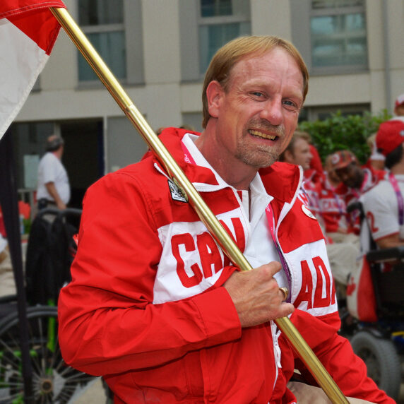 Garett Hickling smiles while proudly holding the Canadian flag, wearing a red and white Team Canada jacket, surrounded by teammates at a Paralympic event.