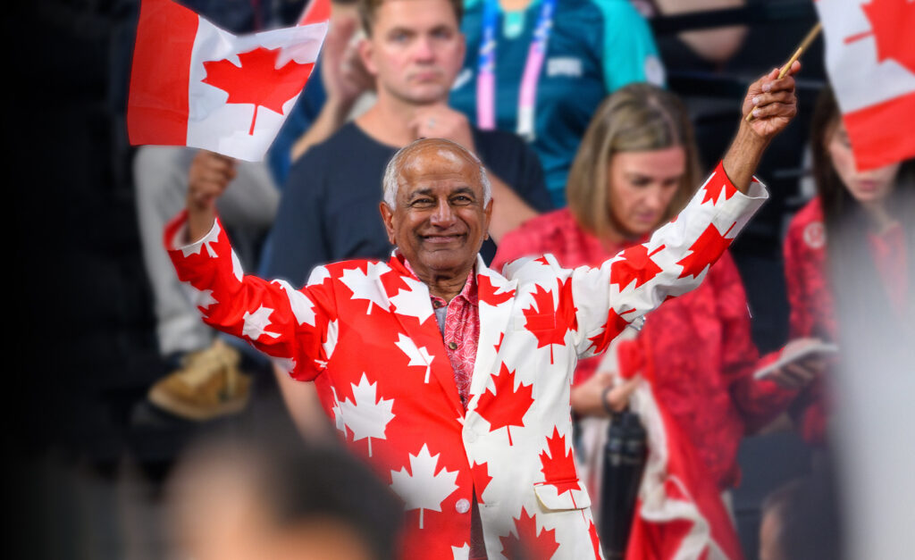 Mike Shaikh, board member of the Paralympic Foundation of Canada, smiles and waves two Canadian flags while wearing a suit covered in red maple leaves at a Canadian Paralympic Team event.