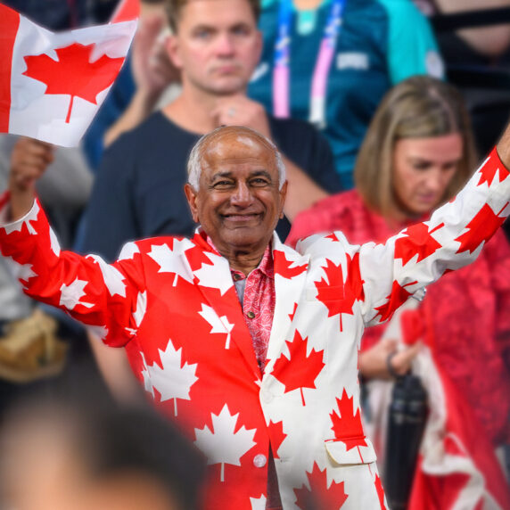 Mike Shaikh, board member of the Paralympic Foundation of Canada, smiles and waves two Canadian flags while wearing a suit covered in red maple leaves at a Canadian Paralympic Team event.