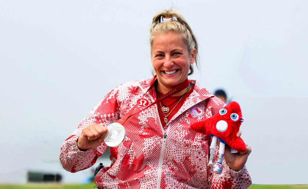 Brianna Hennessy smiles proudly while holding her Paris 2024 silver medal and the red Paralympic mascot plushie, wearing her Team Canada jacket with maple leaf patterns.