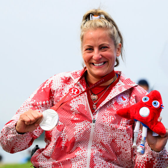 Brianna Hennessy smiles proudly while holding her Paris 2024 silver medal and the red Paralympic mascot plushie, wearing her Team Canada jacket with maple leaf patterns.