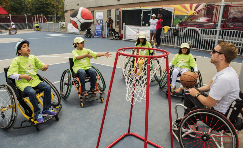 Four children in wheelchairs and an adult play wheelchair basketball outdoors, with the children reaching for a basketball near a hoop.