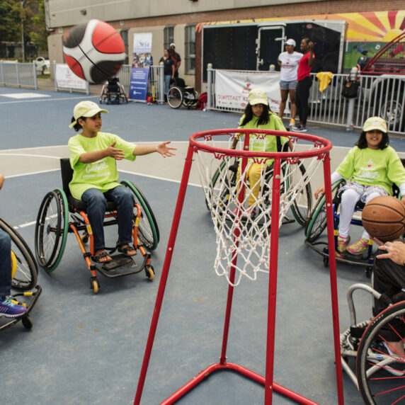 Four children in wheelchairs and an adult play wheelchair basketball outdoors, with the children reaching for a basketball near a hoop.