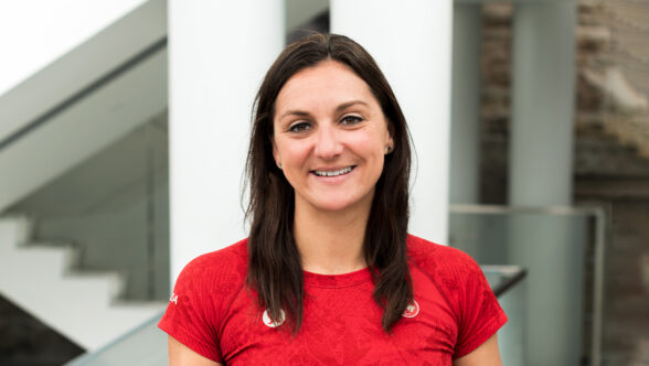 Portrait of Alison Costa smiling, wearing a red Team Canada shirt with two Canadian Paralympic Committee pins, standing in front of a modern white and grey interior background.