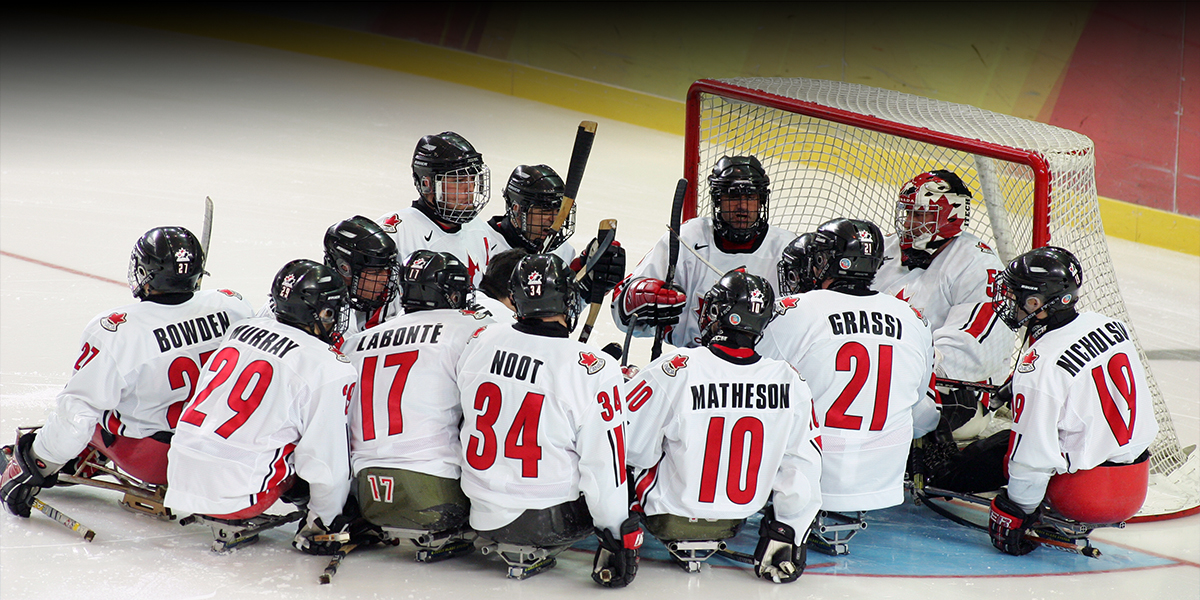 The Canadian Para ice hockey team gathers in a huddle on the ice in front of the goal at the Torino 2006 Paralympic Winter Games. Players wearing white jerseys with red accents listen intently, with some holding their sticks upright.