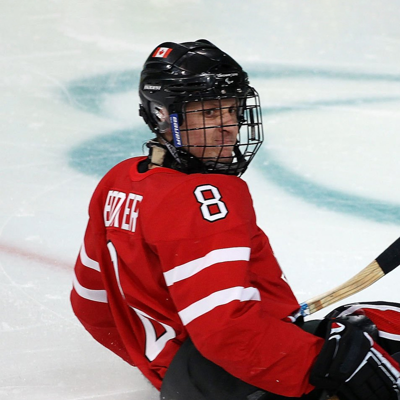 Portrait of Canadian Para ice hockey player Jeremy Booker wearing a red jersey with the number 8, seated in a sled on the ice during a game. He wears a black helmet with a full face cage and a small Canadian flag sticker on top.