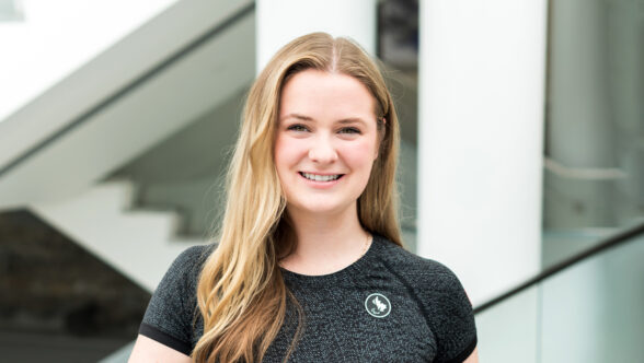 Portrait of Cassidy Cressman smiling, wearing a black athletic Team Canada shirt with a Canadian Paralympic Committee emblem, standing in a modern interior with white columns and a staircase in the background.