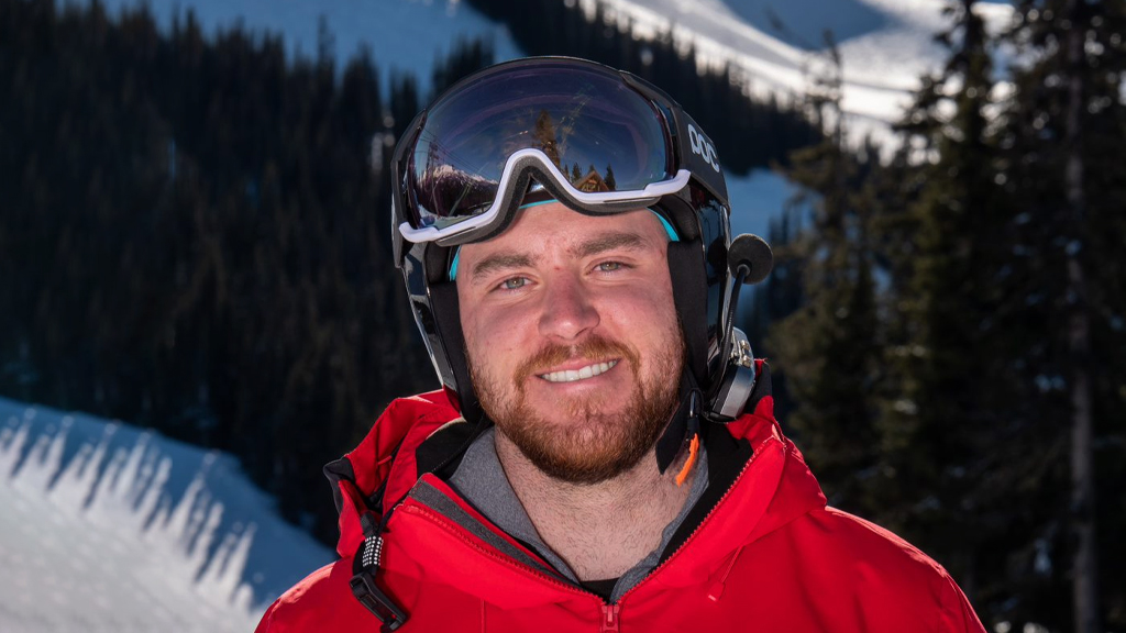 Canadian Paralympian Mac Marcoux smiles while wearing ski goggles and a red jacket, standing on a snowy mountain slope with evergreen trees in the background.