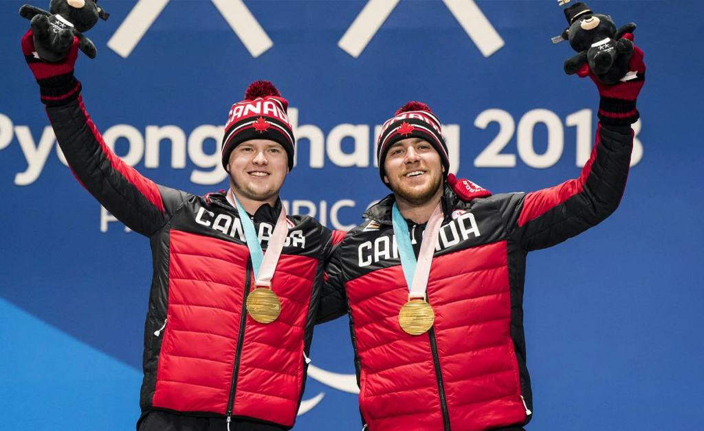 Canadian Para alpine skier Mac Marcoux and his guide stand proudly on the podium at the PyeongChang 2018 Paralympic Winter Games, both holding their arms up in celebration while wearing gold medals and Team Canada jackets and toques.
