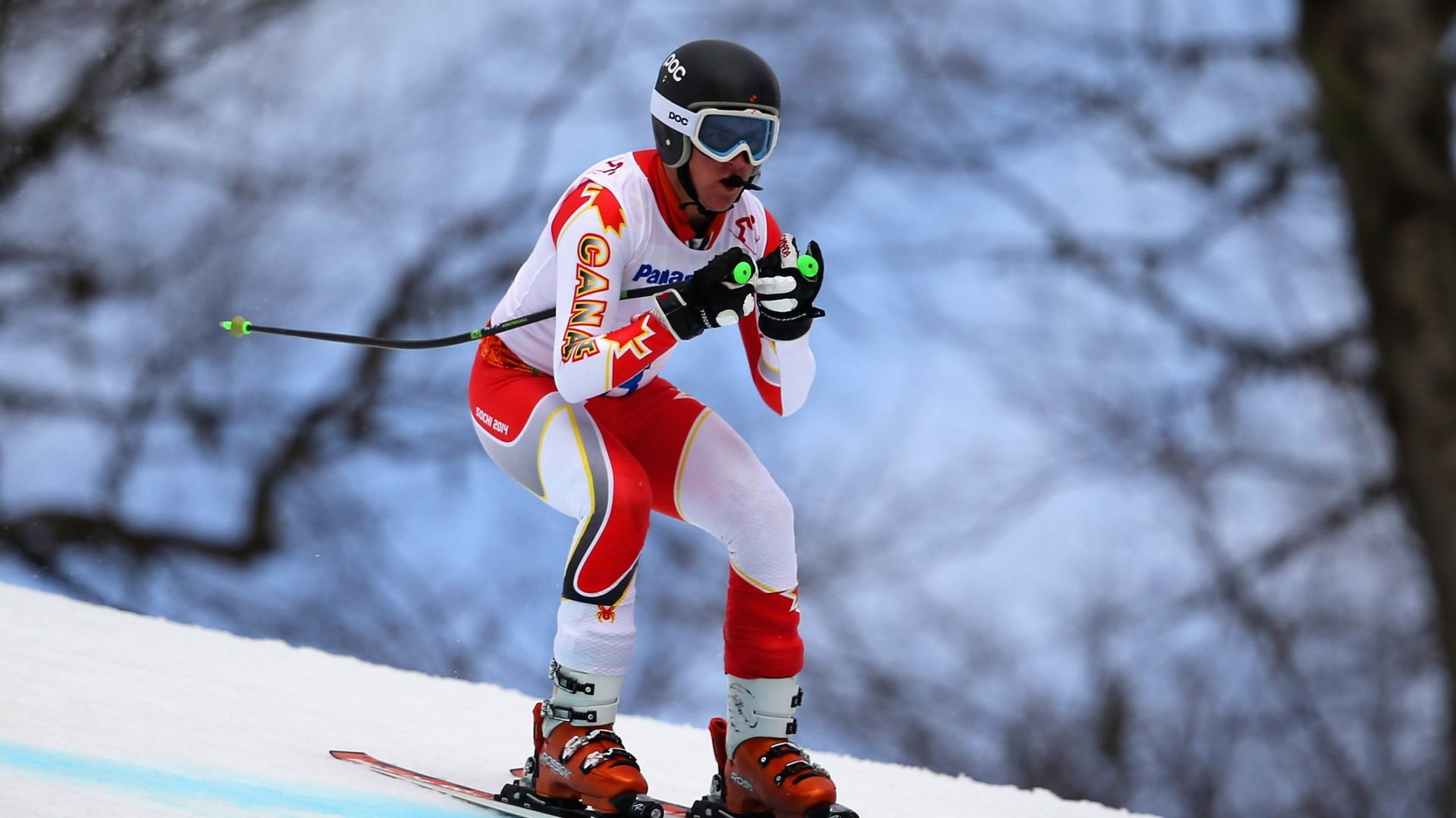 Canadian Para alpine skier Mac Marcoux competes in a downhill race at the Sochi 2014 Paralympic Winter Games, wearing a white and red Team Canada speed suit, helmet, and goggles, in a tucked position on the snowy course.