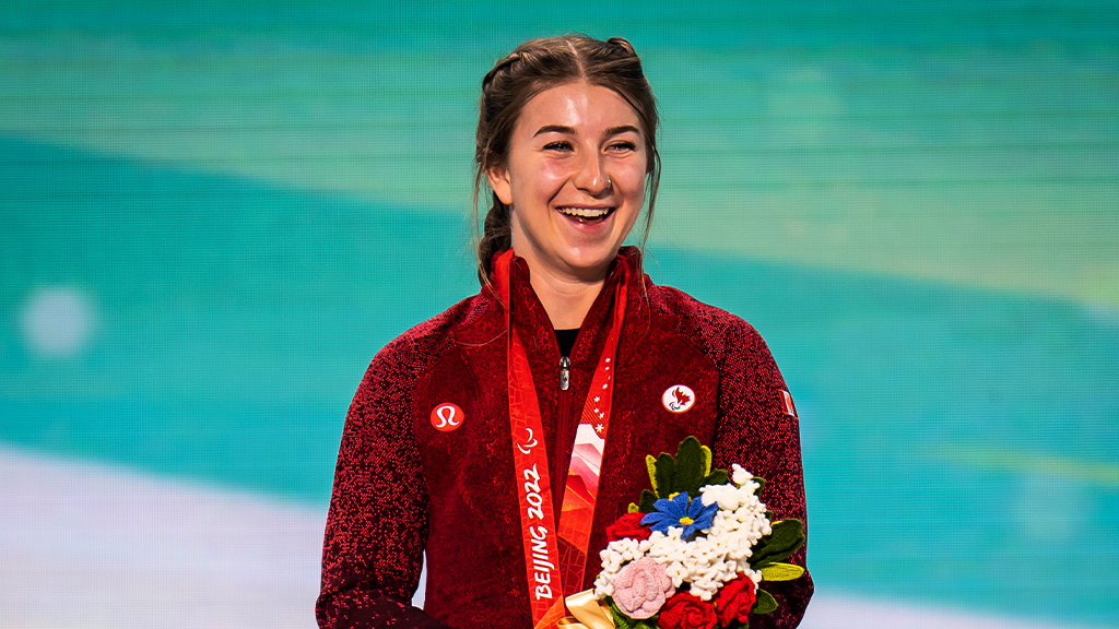 Canadian Para alpine skier Mollie Jepsen smiles on the podium at the Beijing 2022 Paralympic Winter Games, wearing her red Team Canada jacket with a Paralympic medal around her neck and holding a bouquet of victory flowers.