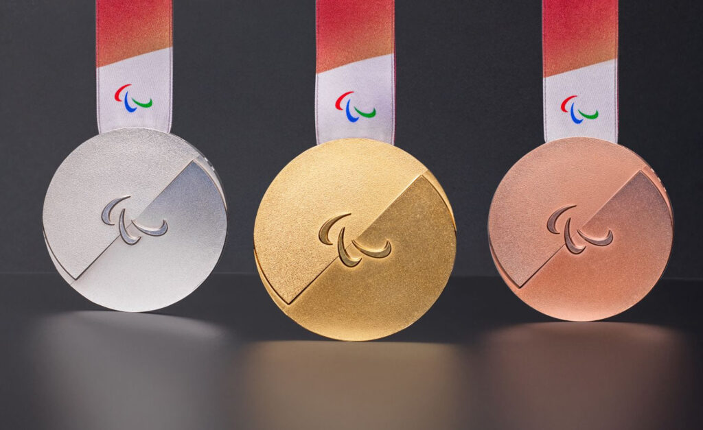 Gold, silver, and bronze medals for the Milano Cortina 2026 Paralympic Winter Games displayed against a dark background. Each medal features a sleek circular design with a diagonal cut and the Paralympic agitos symbol in the centre, with a red-to-white ribbon and embossed Braille.
