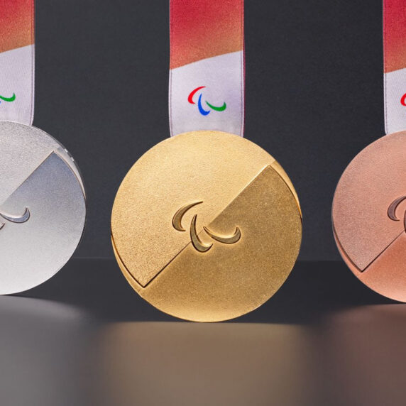 Gold, silver, and bronze medals for the Milano Cortina 2026 Paralympic Winter Games displayed against a dark background. Each medal features a sleek circular design with a diagonal cut and the Paralympic agitos symbol in the centre, with a red-to-white ribbon and embossed Braille.