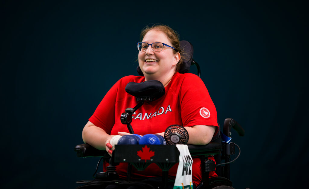 Canadian boccia player Alison Levine smiles while competing at the Santiago 2023 Parapan American Games. She wears a red Team Canada shirt and sits in her power wheelchair with boccia balls and equipment tray in front of her.