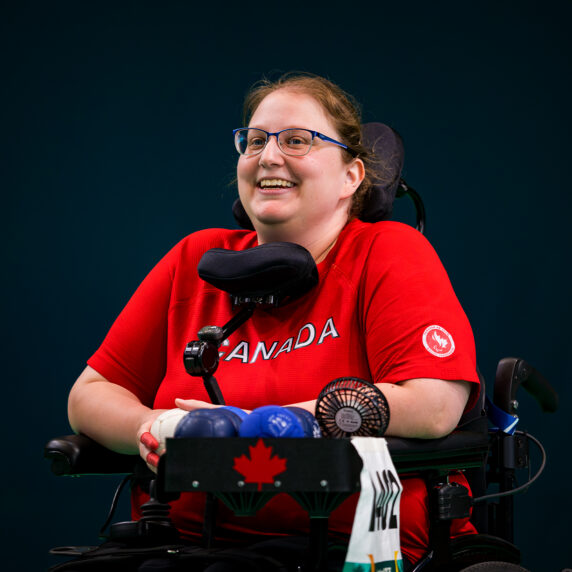 Canadian boccia player Alison Levine smiles while competing at the Santiago 2023 Parapan American Games. She wears a red Team Canada shirt and sits in her power wheelchair with boccia balls and equipment tray in front of her.