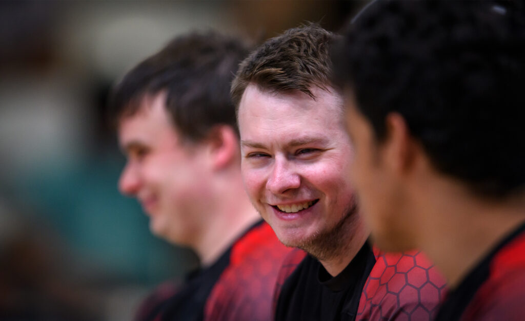 Canadian goalball athlete Blair Nesbitt smiles while sitting on the bench alongside teammates during a match at the Santiago 2023 Parapan American Games. He wears a red and black Team Canada jersey.