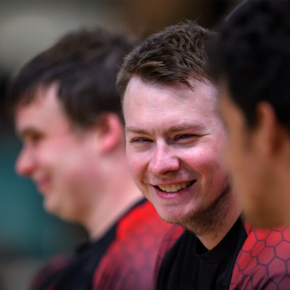 Canadian goalball athlete Blair Nesbitt smiles while sitting on the bench alongside teammates during a match at the Santiago 2023 Parapan American Games. He wears a red and black Team Canada jersey.