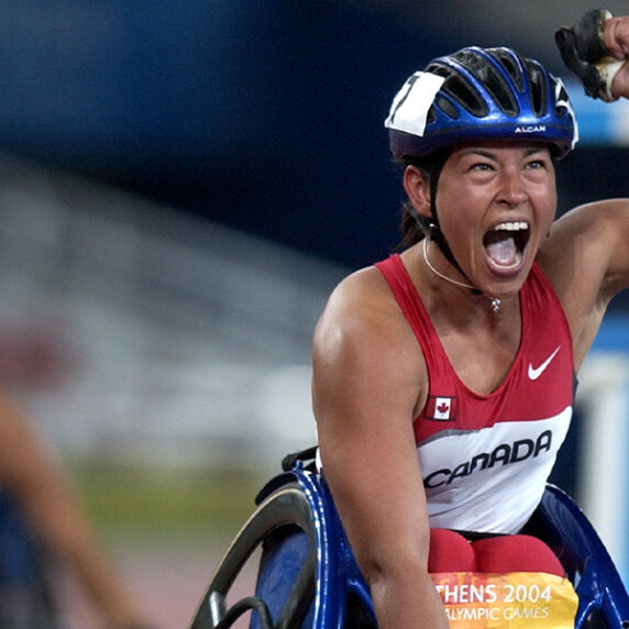 Canadian Paralympian Chantal Petitclerc celebrates with a powerful fist pump after winning a race in wheelchair racing at the Athens 2004 Paralympic Games. Wearing a red and white Team Canada uniform and helmet, she shouts with joy and determination at the finish line.