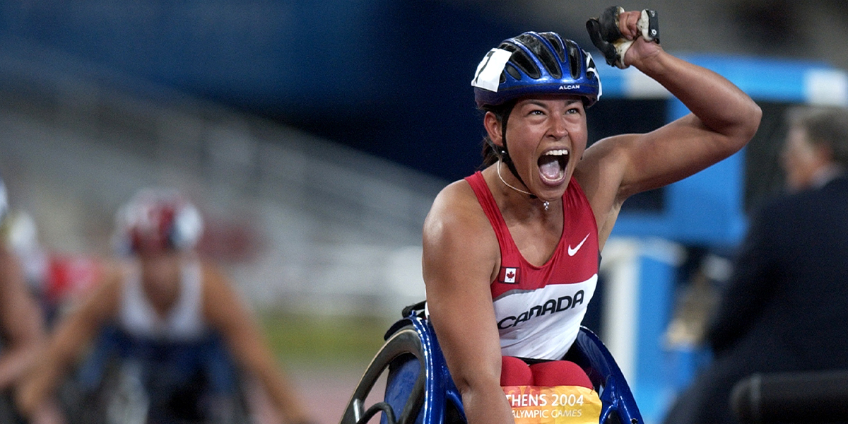 Canadian Paralympian Chantal Petitclerc celebrates with a powerful fist pump after winning a race in wheelchair racing at the Athens 2004 Paralympic Games. Wearing a red and white Team Canada uniform and helmet, she shouts with joy and determination at the finish line.