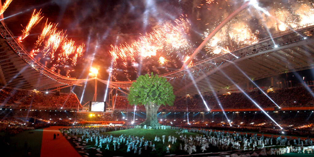 Fireworks light up the night sky above the stadium during the Opening Ceremony of the Athens 2004 Paralympic Games. A large symbolic tree stands in the centre of the stadium, surrounded by performers in white costumes, as spotlights and colourful lights fill the arena.