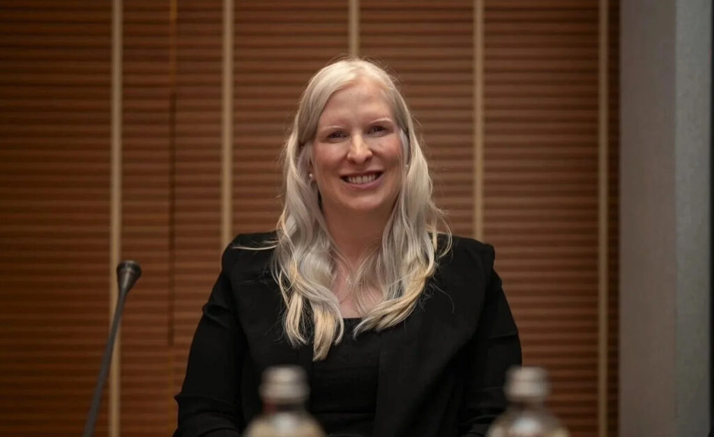 Canadian Paralympian Chelsey Gotell smiles while seated at a conference table, wearing a black blazer with long blonde hair, during an official event.