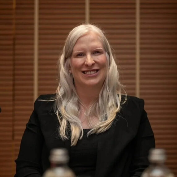 Canadian Paralympian Chelsey Gotell smiles while seated at a conference table, wearing a black blazer with long blonde hair, during an official event.