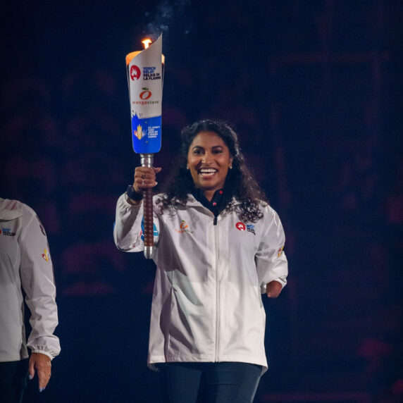 Canadian Paralympian Katarina Roxon smiles while proudly holding the lit torch during the opening ceremony of the 2025 Canada Games. She wears a white Team Newfoundland and Labrador jacket.
