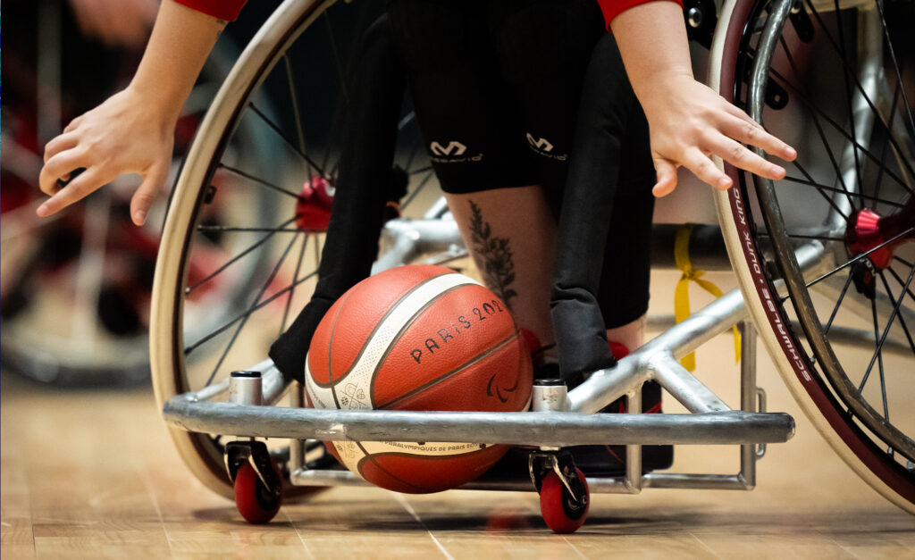 Close-up of a wheelchair basketball game. A player's hand reaches towards the red and white basketball labeled "Paris 2024" on a wooden floor. Dynamic and focused.