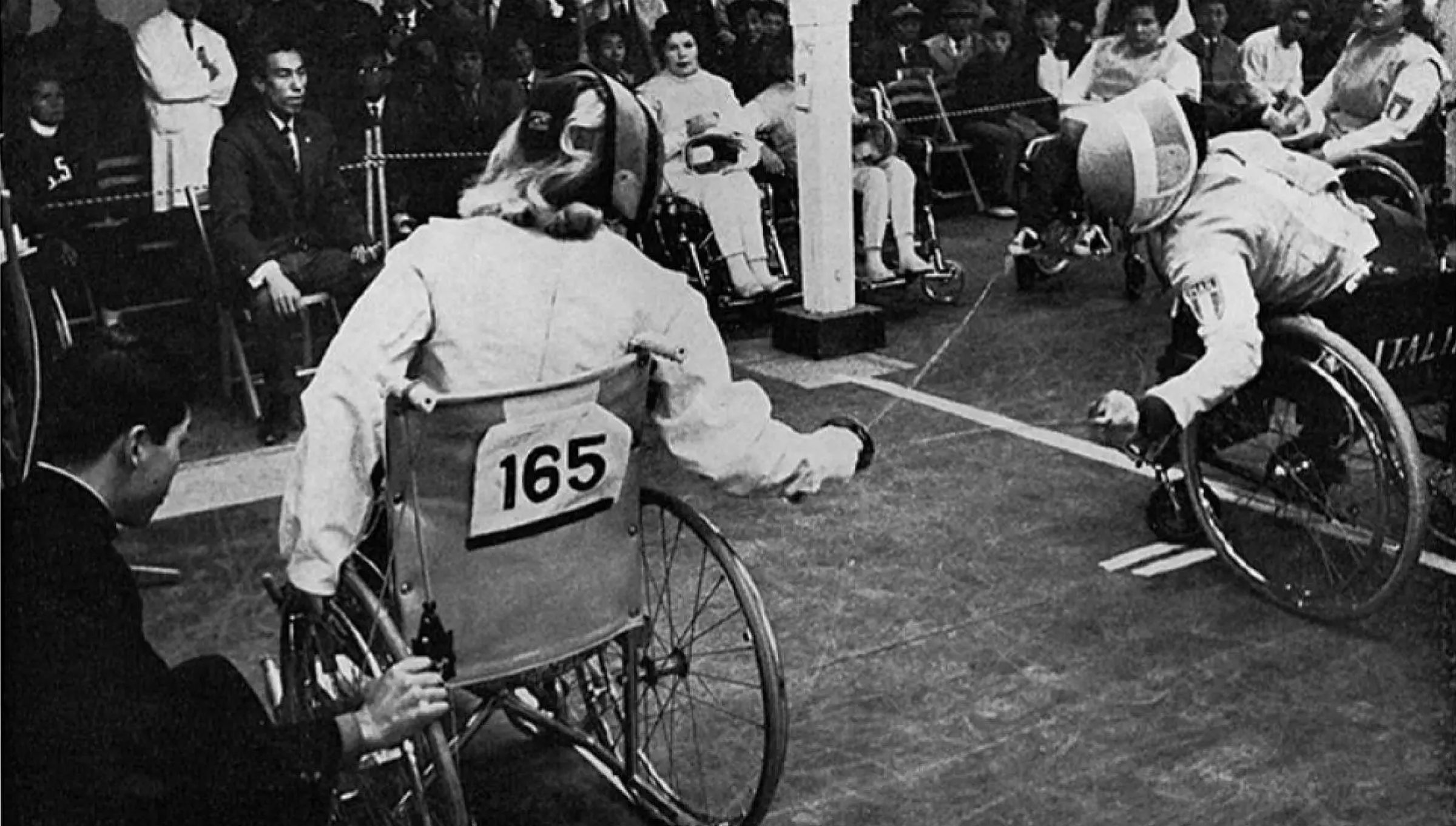 Black and white photo of a Para fencing match at the Tokyo 1964 Paralympic Games. Two wheelchair fencers wearing protective gear face off, with athlete number 165 on the left and an opponent from Italy on the right. A crowd watches in the background.