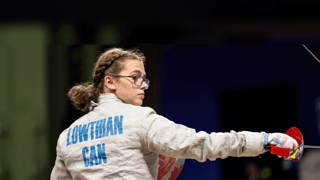Trinity Lowthian wearing glasses and a protective jacket with “LOWTHIAN CAN” on the back extends her arm forward with her foil, focused on her opponent during competition.