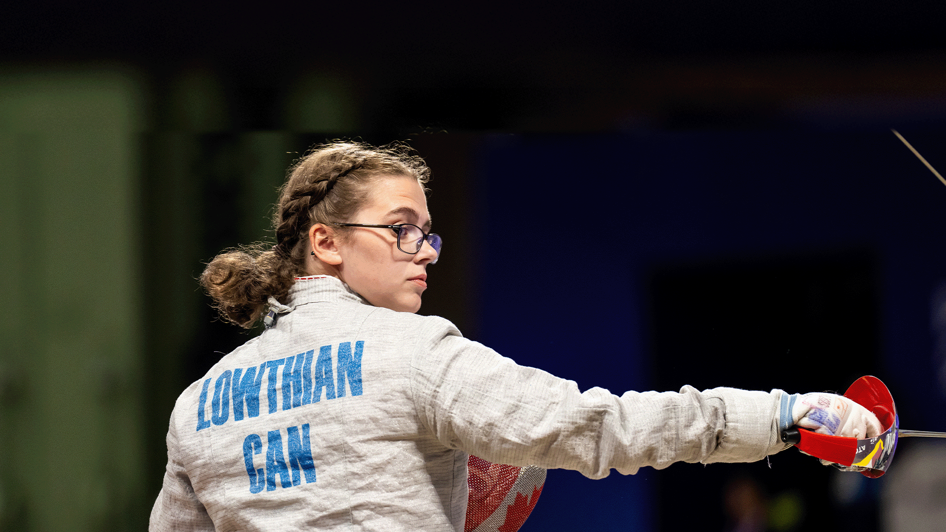 Trinity Lowthian wearing glasses and a protective jacket with “LOWTHIAN CAN” on the back extends her arm forward with her foil, focused on her opponent during competition.